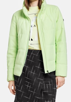 Femme portant une veste matelassée vert clair sur un t-shirt graphique jaune clair et une jupe noire avec un motif à carreaux blanc.