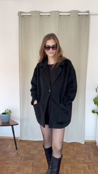 Melns, lielgabarīta, pogu mētelis izgatavots no teksturēta auduma, ar divām priekšējām kabatām, uzvilkts pār melnu kleitu ar fona gaismas aizkariem.