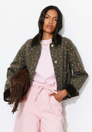 Femme portant une veste fleurie verte, un haut rose et un pantalon rose noué, tenant un sac en daim marron à franges sur fond blanc.