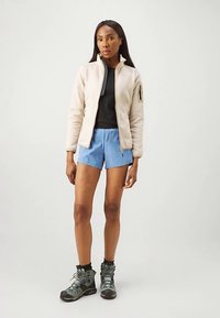 Ζακέτα από φλις σε ανοιχτό μπεζ, μαύρο μακρυμάνικο τοπ, μπλε σορτς και γκρι παπούτσια πεζοπορίας. Μινιμαλιστικός σχεδιασμός με λεπτομέρεια τσέπης με φερμουάρ.