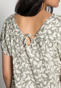 Vrouw met lang, donker haar draagt een groen-witte bloes met bloemenpatroon en een V-hals die op de nek is geknoopt, waardoor haar bovenrug zichtbaar is. Ze draagt gouden oorbellen in de vorm van een ring.