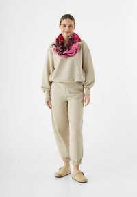Sweatshirt beige et joggers avec des poignets élastiques, associés à une écharpe à motifs rose et bordeaux. Tissu doux, coupe décontractée.