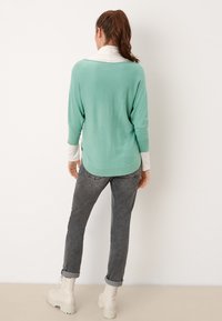 Pull vert menthe à manches longues superposé à une chemise blanche à col, associé à un jean gris et des bottines de couleur claire.