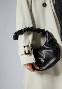 Sac en cuir noir avec une bandoulière froncée et un détail logo doré, porté par une personne vêtue d'un manteau clair à boutons.