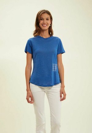 T-shirt bleu à manches courtes en tissu doux, avec un ourlet incurvé et un détail brodé blanc sur le côté inférieur gauche.
