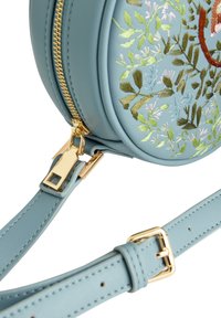 Borsa rotonda in pelle azzurro chiaro con ricamo floreale, accessori in metallo dorato sulla zip e una tracolla regolabile in pelle coordinata. Texture liscia.