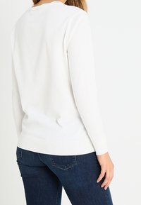 Valkoinen pitkähihainen neule pyöreällä kaula-aukolla ja resorilla helman kohdalla. Pehmeä tekstuuri ja istuva malli, yhdistetty tummansinisiin farkkuihin.