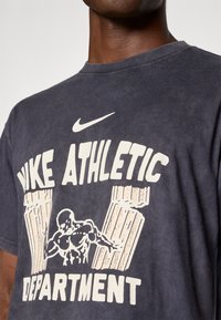 Tamsiai mėlynas medvilninis marškinėliai su baltu grafiką, kuriame parašyta "Nike Athletic Department" ir iliustracija raumeningo figūros.