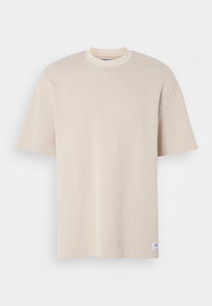 Beige ribbed t-shirt with a round neckline and short sleeves, featuring a visible brand label at the bottom left corner.