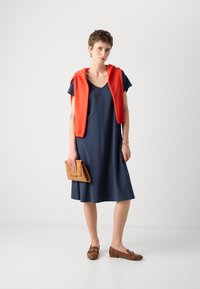 Navy blue short-sleeve dress with a V-neck, paired with a red sweater draped over the shoulders, brown loafers, and a tan clutch.