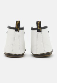 Botas de tobillo de cuero blanco con ribetes negros, acentos de costura amarilla y tiradores con texto negro y amarillo en la parte posterior.
