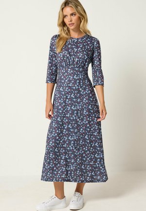Donna che indossa un vestito midi blu navy a fiori con maniche a tre quarti e scarpe da ginnastica bianche, in piedi davanti a uno sfondo semplice.