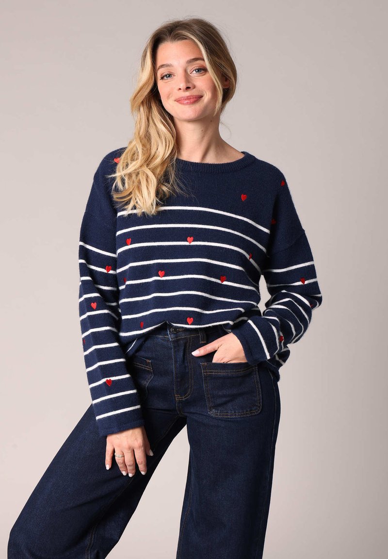 Pull marin rayé avec des accents de cœur rouge, texture en tricot, manches larges et encolure ronde, associé à un pantalon taille haute en denim foncé.