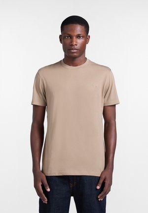 Muž stojaci čelom dopredu, oblečený v béžovom tričku s krátkym rukávom a okrúhlym výstrihom a tmavomodrých džínsoch na bielom jednoduchom pozadí.