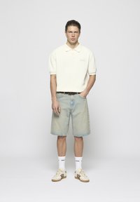Camisa polo blanca de manga corta, pantalones cortos de mezclilla azul claro, zapatillas beige con acentos marrones, calcetines blancos con texto negro.