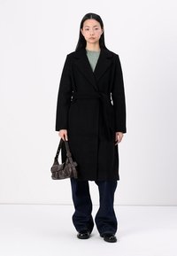 Manteau long noir avec ceinture, col à revers, et texture lisse, associé à un haut en maille verte et un jean large bleu marine. Tenant un sac marron.