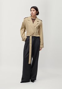 Beige wikkeljas met een riem, brede zwarte broek, effen kleuren, gladde textuur en dubbelgebreid ontwerp; model voor een neutrale achtergrond.