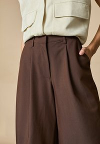 Brune, højtaljede bukser med en glat tekstur, plisseret design, sidelommer og en afslappet pasform, parret med en lys beige top.