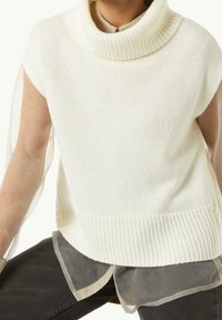 Persoon in een mouwloze witte gebreide coltrui over een doorschijnende blouse met patroon en lange mouwen, met donkere broek, zittend.