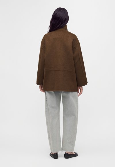 Femme aux longs cheveux foncés, debout de dos, portant un manteau marron, un jean gris clair ample et des chaussures plates noires sur un fond uni.