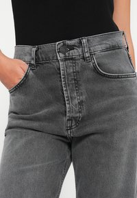 Jeans en denim gris avec une taille haute, comprenant cinq poches, une fermeture à bouton en métal et des détails de couture visibles.