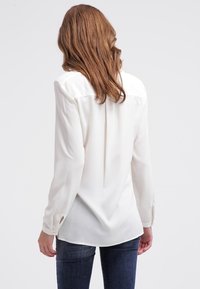 Langærmet bluse i hvid silke, med en knappet manchet, rygfald detaljer og en glat tekstur. Kombineret med mørkeblå jeans.