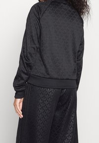 Svart idrottströja med tonalt lövmönster, ribbade ärmslut och fåll. Stylad med matchande svarta byxor som har liknande designelement.