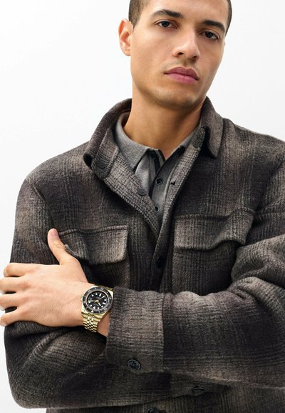 Jeune homme portant une veste à carreaux sombre et une chemise grise, les bras croisés, montrant une montre-bracelet dorée avec un cadran noir.
