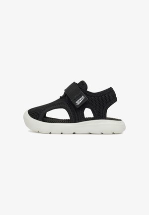 Sandale en mesh noir respirant avec semelle blanche flexible et bride Velcro ajustable sur fond blanc.