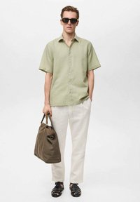 Camicia a maniche corte, verde chiaro, con bottoni, pantaloni crema, sandali neri e una borsa marrone. Il modello indossa occhiali da sole scuri. Sfondo semplice.