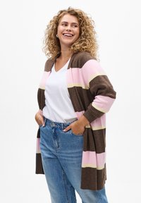 Cardigan marron avec des rayures roses et jaunes, devant ouvert, manches longues et poignets côtelés. Porté par-dessus un haut blanc à col en V et un jean bleu.