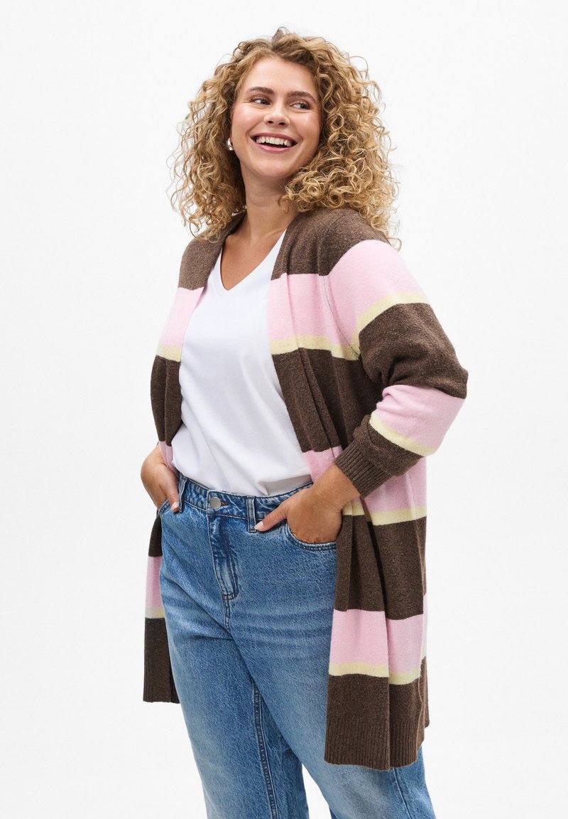 Cardigan marron avec des rayures roses et jaunes, devant ouvert, manches longues et poignets côtelés. Porté par-dessus un haut blanc à col en V et un jean bleu.