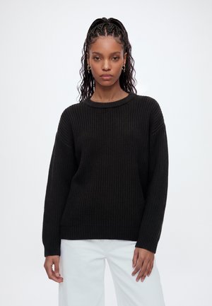 Jeune femme aux cheveux tressés portant des boucles d'oreilles créoles dorées, un pull en maille noir et un pantalon blanc sur un fond blanc uni.