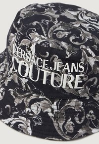 Čierny klobúk s pokrývkou hlavy s ozdobnými sivými kvetinovými vzormi, na ktorých sú v tučných bielych písmenách napísané slová "VERSACE JEANS COUTURE". Bavlnený materiál.