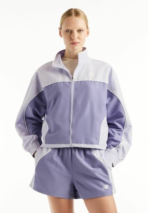 Ung person iført en lyse-lilla og lavendelfarvet jakke med lynlås og matchende shorts, stående med hænderne i lommerne mod en hvid baggrund.