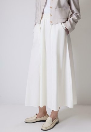 Persona che indossa una giacca corta beige, pantaloni bianchi a gamba larga a vita alta e mocassini beige, in piedi contro un muro semplice.