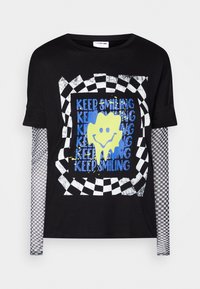 T-shirt nera a maniche corte stratificata con maniche lunghe a quadri, caratterizzata da una grafica centrale di un volto sorridente e testo ripetuto in blu e giallo.