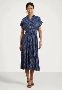 Sieviete, kas tērpusies zilā rakstainā midi apģērbā ar īsām piedurknēm un brūnām potītes siksnām, stāvot pret baltu fonu.