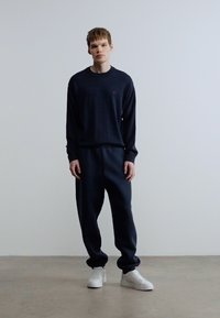 Ναυτικό μπλε πουλόβερ και τζόγκερς σε απαλό ύφασμα, με ελαφρώς χαλαρή εφαρμογή. Στυλιζαρισμένα με λευκά αθλητικά παπούτσια. Μινιμαλιστικός σχεδιασμός, χωρίς σχέδια.