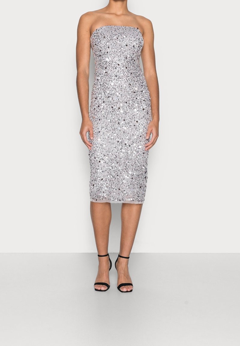 Robe midi argentée sans bretelles à sequins, cintrée à la taille, portée avec des talons ouverts à bride cheville noirs sur un fond uni.
