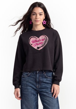 Ung kvinde med langt mørkt hår iført sort sweatshirt med lyserødt hjerte og teksten "What's up Pookie", stjerneformede lyserøde øreringe og blå jeans.