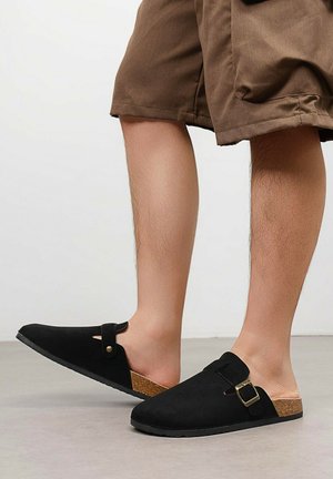 Jambes portant un short marron et des sabots noirs sans lacets avec des brides à boucle et une semelle en liège, debout sur un sol gris contre un mur blanc.