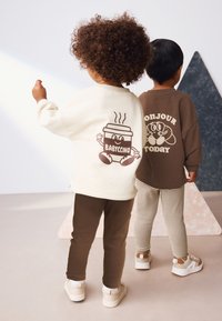 To børn står ryg mod ryg, iført en cremefarvet sweatshirt med et kaffe-motiv og en brun sweatshirt med en tegneserie. Brune og stribede bukser.