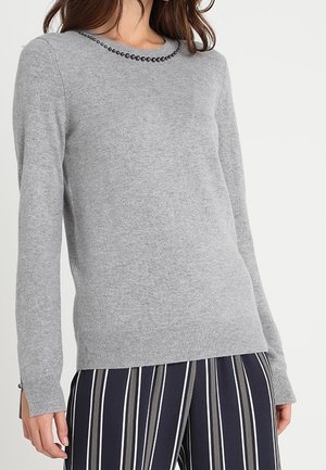 Mujer con un suéter de manga larga gris claro con detalles de cuentas negras en el cuello y pantalones azul marino con rayas blancas verticales.