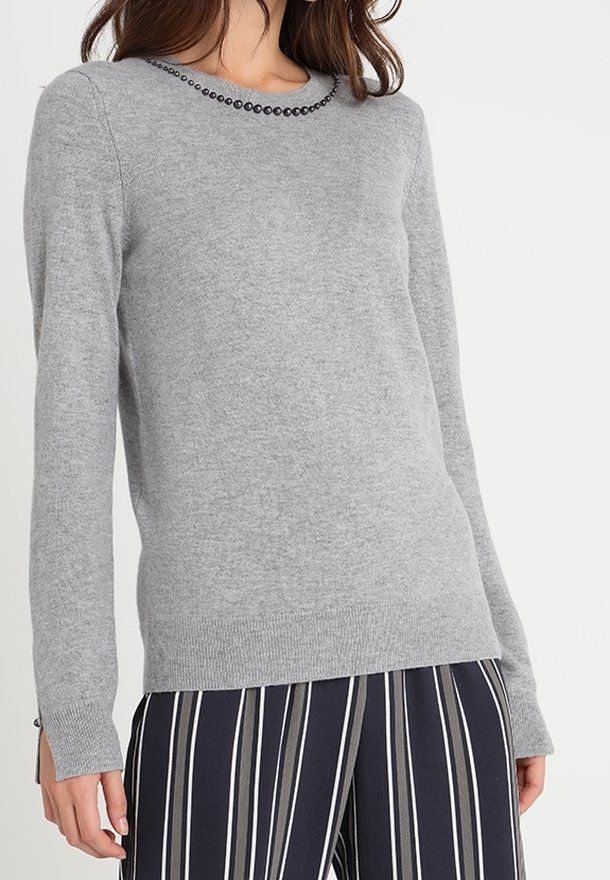 Femme portant un pull léger gris clair à manches longues avec des perles noires au col et un pantalon bleu marine à rayures verticales blanches.