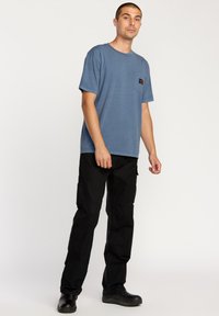 Blå kortærmet t-shirt med brystlomme, kombineret med sorte cargobukser. Stoffet ser blødt ud, med en afslappet pasform og ingen synlige mønstre.