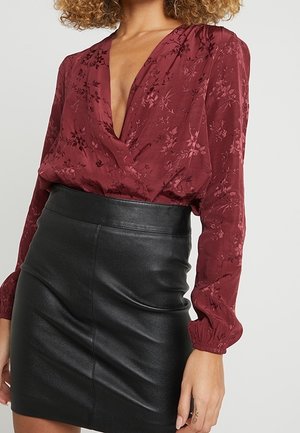 Femme portant un chemisier à fleurs rouge profond à manches longues avec un décolleté plongeant, rentré dans une mini-jupe en cuir noir taille haute.