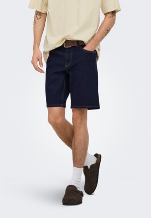 Henkilö, jolla on beige paita, tummat denimshortsit, ruskea vyö, valkoiset sukat ja ruskeat pistokkaat, toinen käsi taskussa, kuvattuna olkapäistä alaspäin.