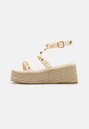 Beige plateauzool sandaal met gevlochten touwzool, gouden piramidevormige studs op de bandjes en verstelbare enkelgesp op een witte achtergrond.