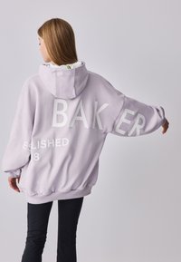 Lys lilla oversized hættetrøje med stor hvid tekst "BAKER" og "ESTABLISHED 88" på ryggen; har hætte og elastiske manchetter.